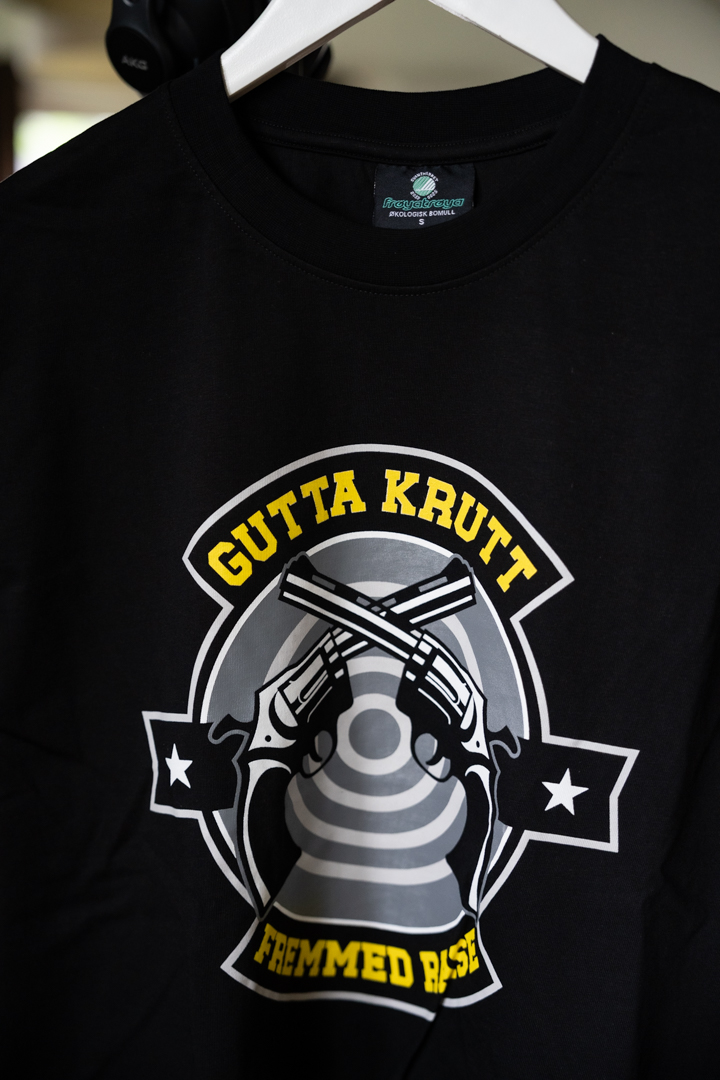 Svart t-skjorte med “Gutta Krutt”-motiv i offwhite, støvet gul, kullgrå og militærgrå toner.